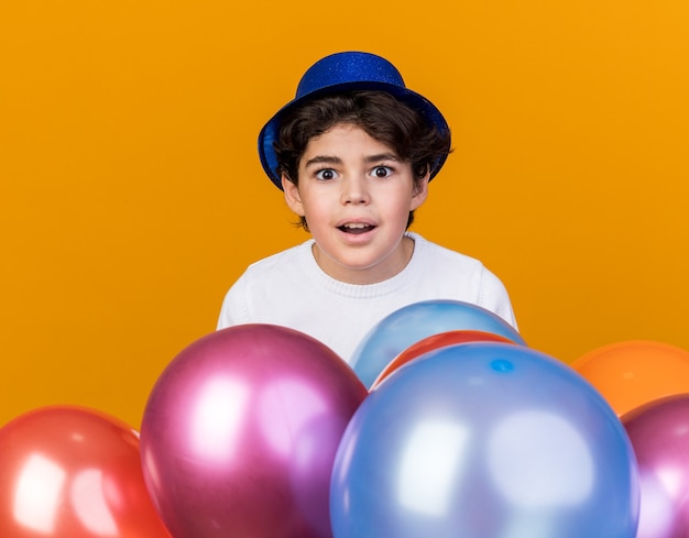 Niño sorprendido con sombrero de fiesta azul de pie detrás de globos aislados en la pared naranja