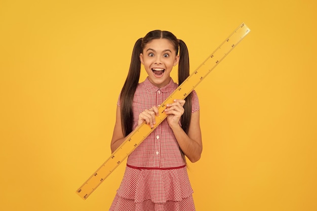 Niño sorprendido sostenga la regla de la herramienta de matemáticas de la escuela en la escuela de fondo amarillo