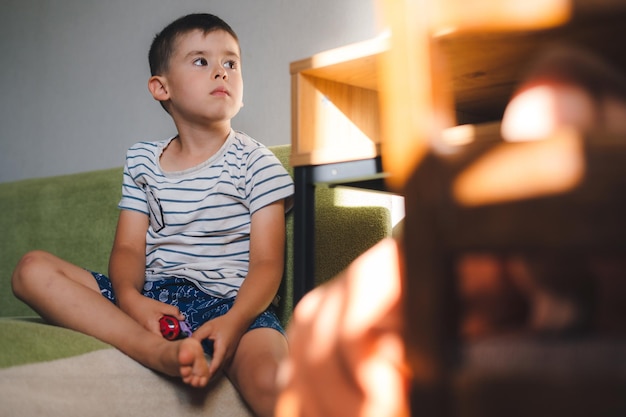 Niño sosteniendo algunos juguetes en sus manos y sentado en el sofá mientras guiña un ojo a la caricatura que está en la televisión Ocio familiar Interior de la habitación