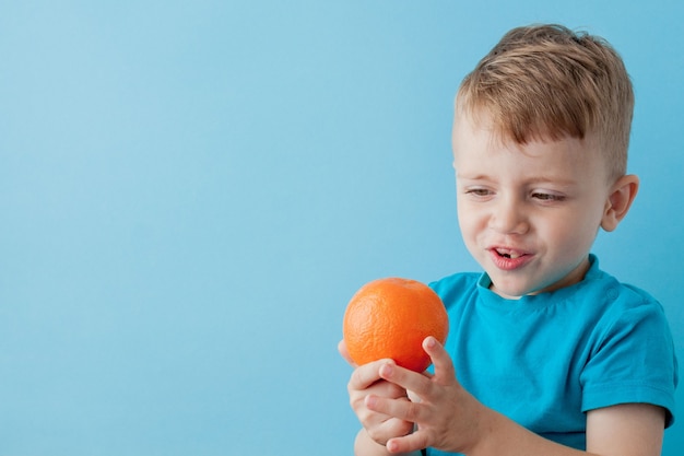 Niño sosteniendo naranja en sus manos sobre la pared azul, dieta y ejercicio concepto de buena salud