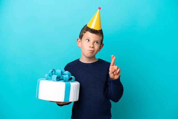 Niño sosteniendo pastel de cumpleaños aislado de fondo azul con los dedos cruzados y deseando lo mejor