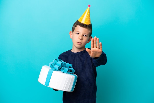 Niño sosteniendo pastel de cumpleaños aislado sobre fondo azul haciendo gesto de parada
