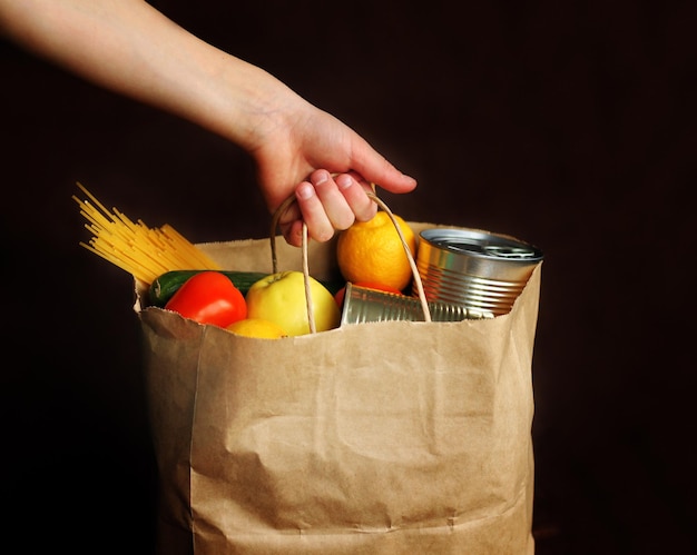 Niño sostiene una bolsa de papel con productos sobre un fondo marrón. Un niño toma comida del servicio de entrega de comida. Alimentos enlatados, pastas, verduras, frutas. Entrega sin contacto.