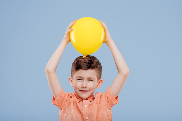 Foto niño sostiene un globo amarillo. mira a la cámara, aislada sobre fondo azul, espacio de copia