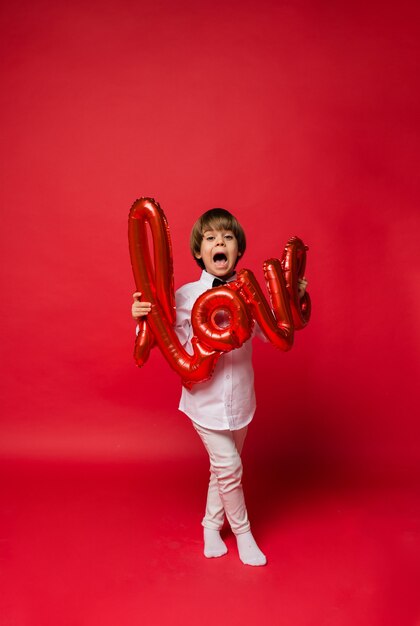 niño se para y sostiene un globo de amor rojo sobre un fondo rojo con espacio para texto