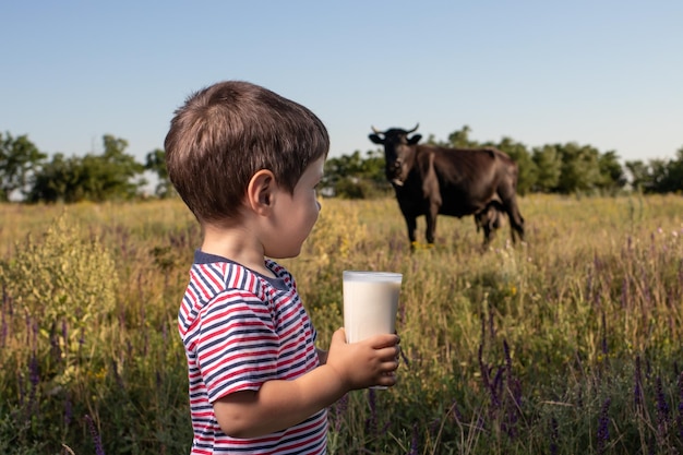 Un niño sostiene un vaso de leche en un campo cerca de una vaca.