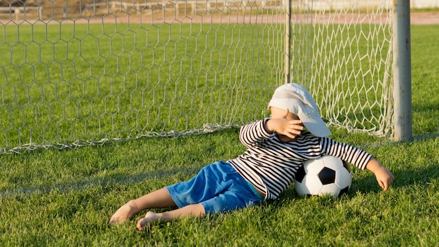 Niño con su balón de fútbol descalzo sobre la hierba en las metas protegiéndose los ojos contra el resplandor del sol vespertino