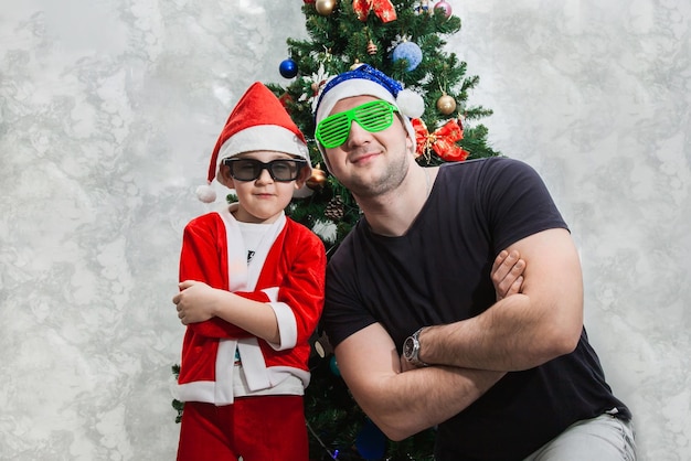 Un niño y su padre con gafas de sol divertidas y gorros de Papá Noel están parados cerca del árbol de Navidad con los brazos cruzados sobre el pecho Juegos para Navidad con la familia