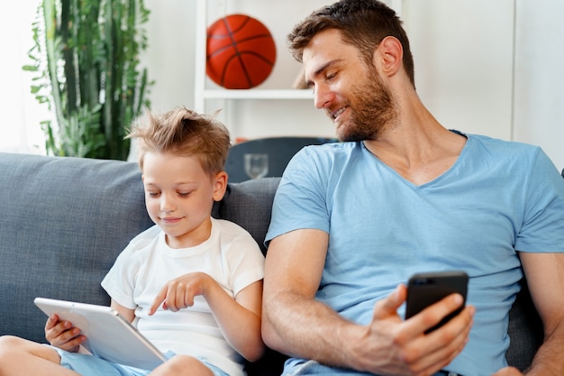 Niño y su papá viendo videos en tableta digital juntos sentados en el sofá en casa