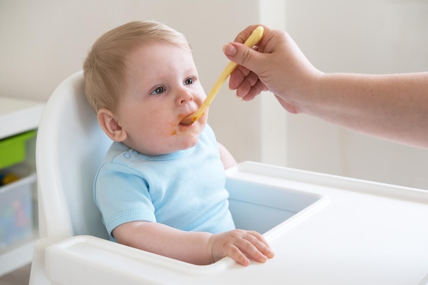 Niño sucio comiendo comida triturada blanda sentado en una silla alta madre alimentando al niño