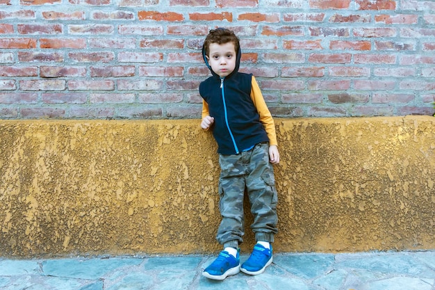 Niño con sudadera con capucha y pantalones de camuflaje apoyado contra la pared de ladrillo