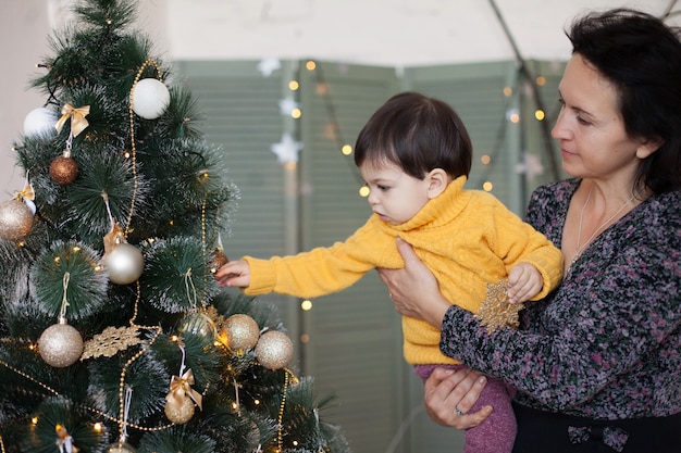 Un niño con un suéter amarillo alcanza una pelota en un árbol de Navidad sentado en los brazos de mamá.