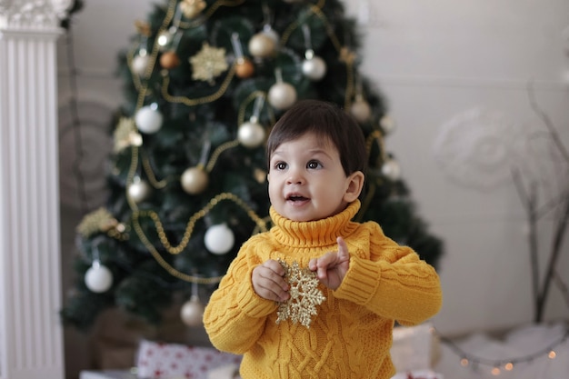 Foto niño con un suéter amarillo sostiene una estrella dorada de navidad en sus manos y sonríe cerca del árbol de navidad y una chimenea.