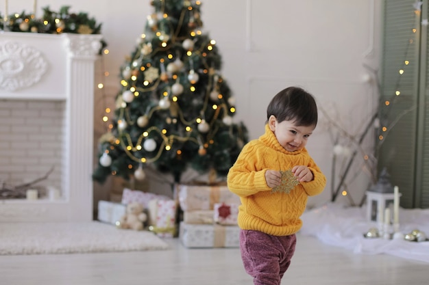 Niño con un suéter amarillo sostiene una estrella dorada de Navidad en sus manos y sonríe cerca del árbol de Navidad y una chimenea.