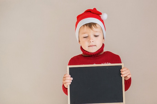 Niño con suéter de Navidad y gorro de Papá Noel con pizarra.