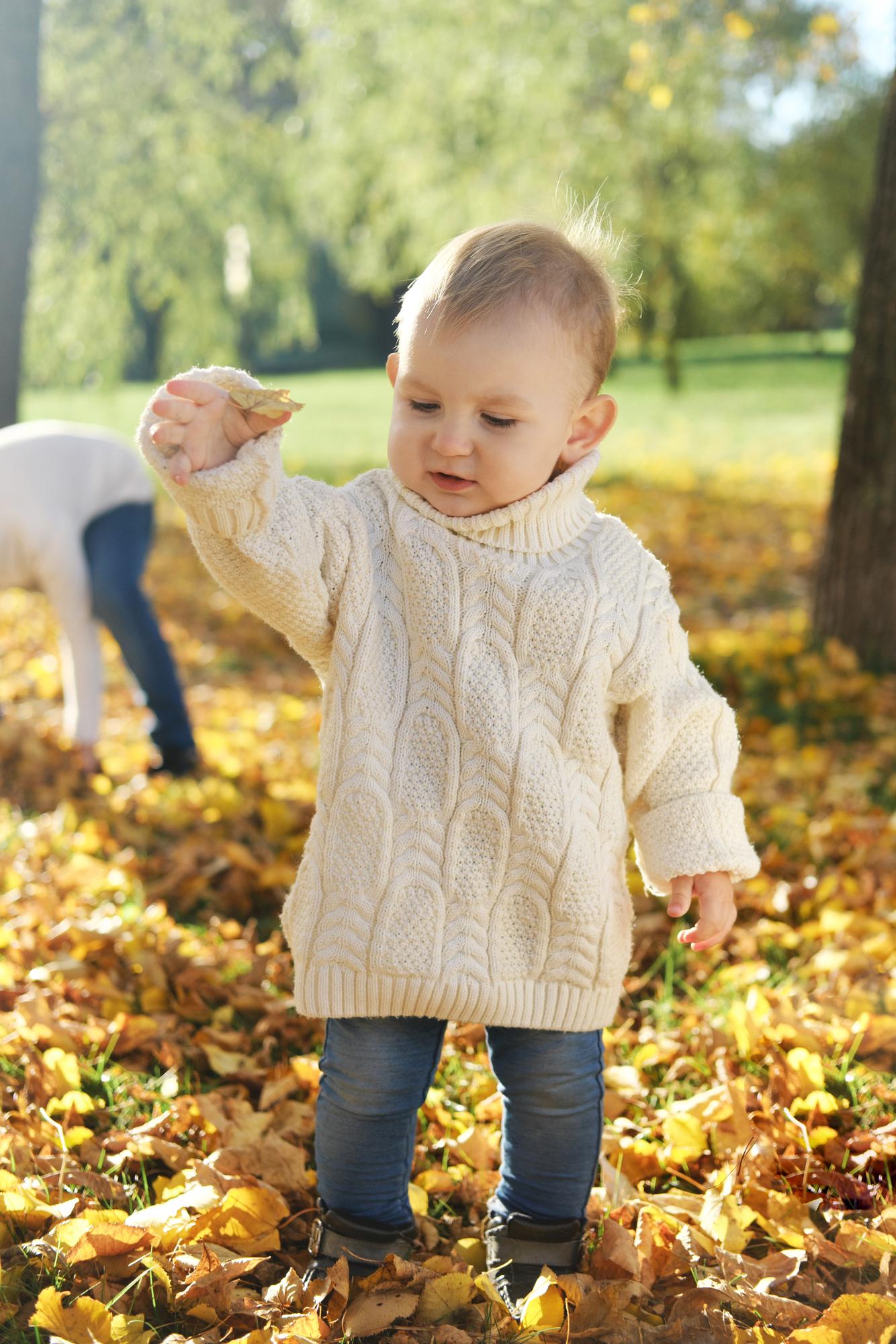 Un niño con un suéter de punto blanco juega con hojas de otoño en el parque Foto vertical