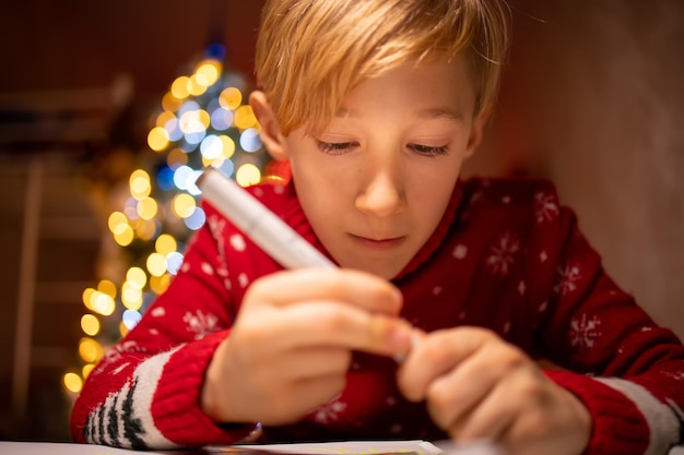 Un niño con un suéter rojo de Navidad en el fondo de un árbol de Navidad se dedica al proceso de dibujo en su habitación bajo una lámpara