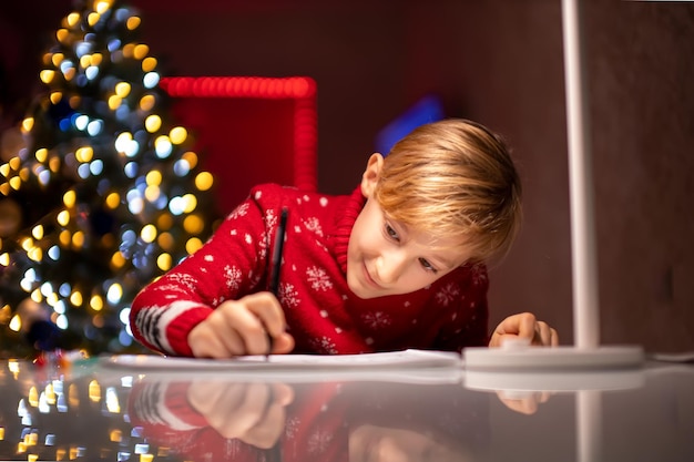Un niño con un suéter rojo de Navidad en el fondo de un árbol de Navidad dibuja algo en su habitación