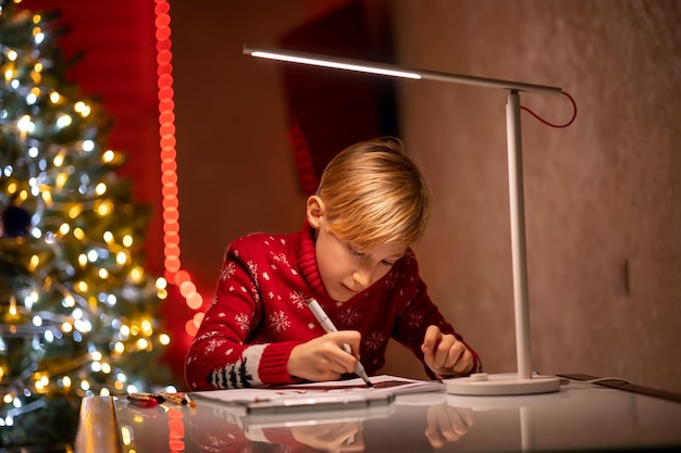 Un niño con un suéter rojo de Navidad en el fondo de un árbol de Navidad iluminado por una lámpara sostiene un marcador de dibujo en la mano