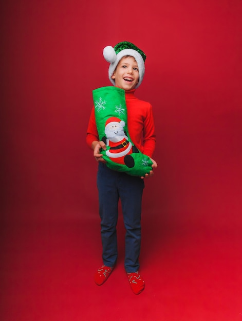 Un niño con un suéter rojo de punto y un sombrero de Papá Noel sostiene una bota de Navidad para regalos Un bebé lindo está esperando la Navidad y un nuevo tipo de aislamiento en un fondo rojo