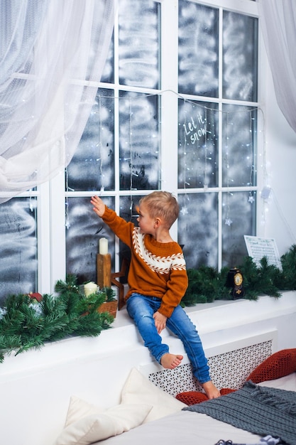 Niño en un suéter se sienta en el alféizar de la ventana en casa esperando milagros regalos y Navidad
