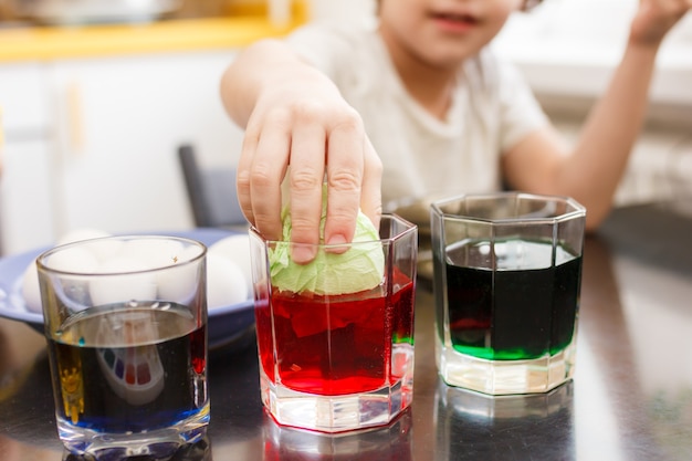 Foto niño sumerge un huevo en un vaso con pintura. niño pinta huevos de pascua.