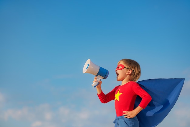 Niño superhéroe gritando a través del altavoz al aire libre. niño súper héroe contra el fondo azul del cielo de verano. concepto de imaginación y libertad.