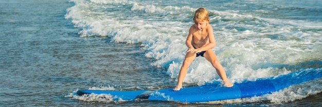 Niño surfeando en una playa tropical. Niño en tabla de surf en ola oceánica. Deportes acuáticos activos para niños. Niño nadando con surf. Lección de surf para niños. BANNER, FORMATO LARGO