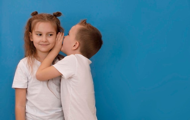 Foto el niño susurra al oído de la niña diciéndole algo secreto buenas noticias sobre fondo azul con un espacio vacío para copiar