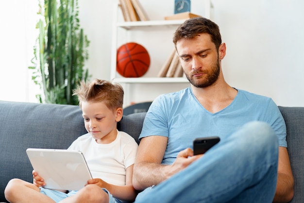 Niño con tableta digital y su padre con smartphone se sientan juntos en el sofá en casa