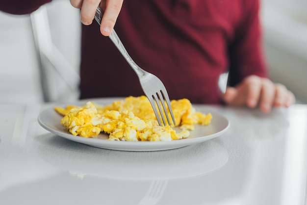 Un niño por la tarde en una cocina de luz blanca con un suéter burdeos come una tortilla