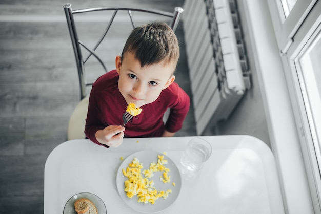 Un niño por la tarde en una cocina de luz blanca con un suéter burdeos come una tortilla