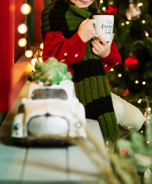 Un niño con una taza de bebida caliente en sus manos se sienta en el fondo de un árbol de Navidad, en primer plano hay un carro de juguete. Foto