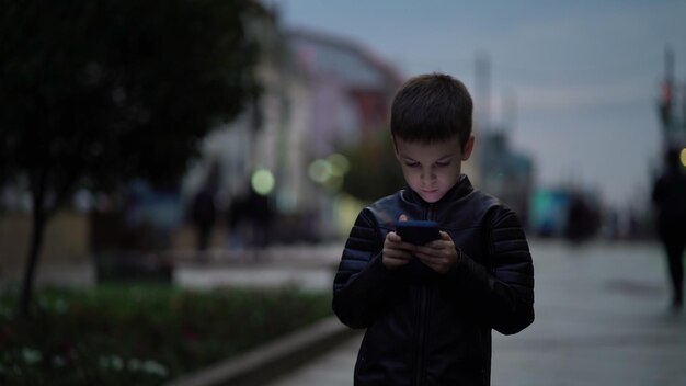 Foto niño con un teléfono inteligente moderno camina por la ciudad nocturna a lo largo de la calle