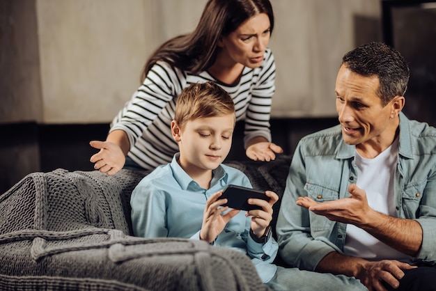 Niño terco. Un niño preadolescente de pelo rubio jugando con el teléfono serenamente, sin prestar atención a que sus padres lo regañen y le pidan que cuelgue el teléfono.