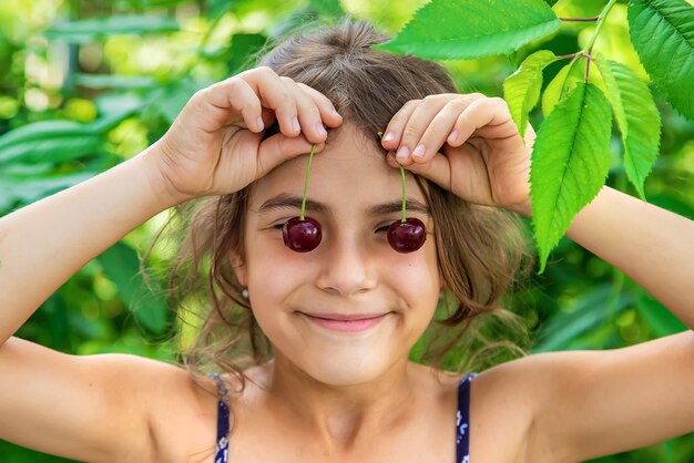 El niño tiene cerezas sobre sus ojos en el fondo del jardín