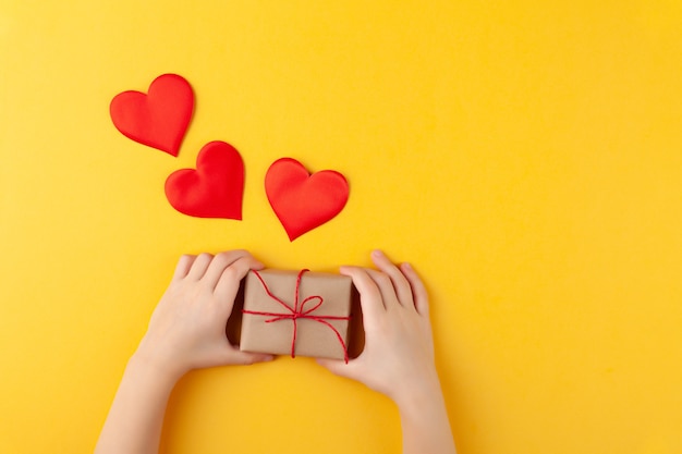 El niño tiene en las manos una caja de regalo sorpresa, muchos corazones rojos, el amor y el concepto del día de San Valentín, horizontal, pared amarilla, espacio de copia, vista superior, endecha plana