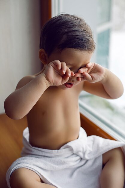 Foto niño en toalla de baño después de lavarse sentado en el alféizar de la ventana en la ventana grande en invierno