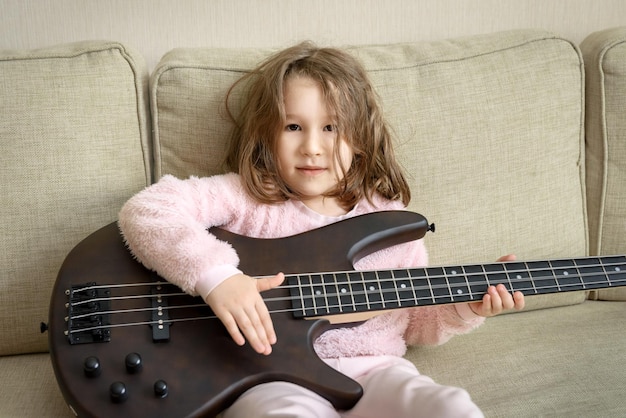 Niño tocando el bajo en casa retrato de una niña pequeña sentada en el sofá con un instrumento musical feliz y lindo niño golpea las cuerdas de la guitarra