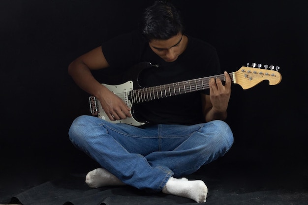 Niño tocando la guitarra eléctrica en un estudio negro sentado con las piernas cruzadas en el suelo