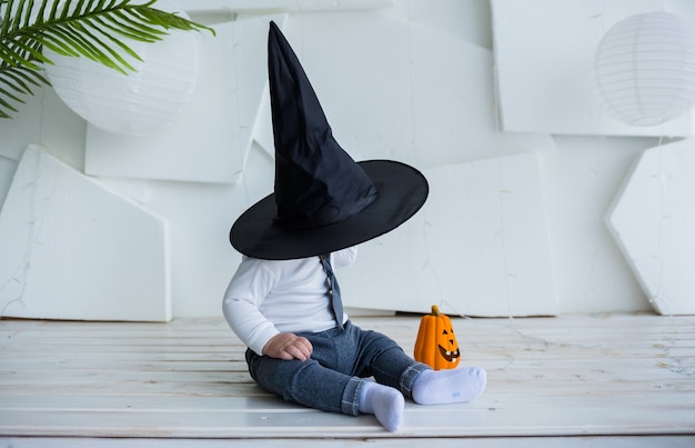 Un niño con un traje se cubrió con un sombrero negro y se sienta con una calabaza sobre un fondo blanco con un lugar para el texto.
