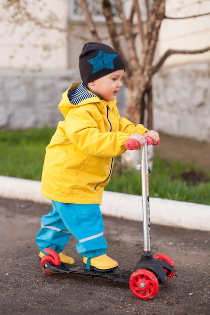 Foto un niño con un traje protector de agua monta un scooter a través de charcos. paseos de primavera al aire libre