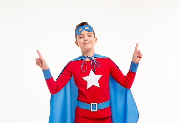 Foto niño en traje de superhéroe brillante mirando a la cámara con una sonrisa y apuntando hacia arriba contra el fondo blanco.