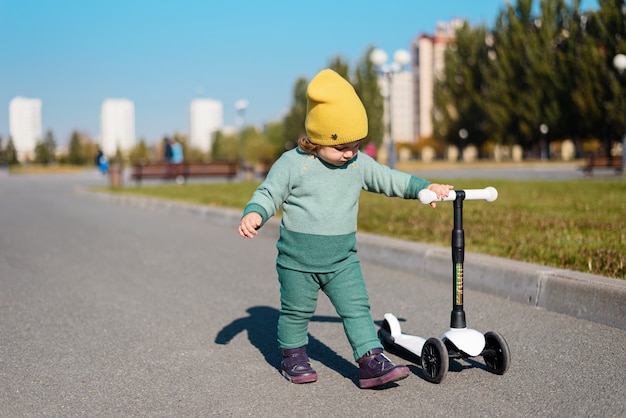 Niño en traje verde, sombrero amarillo monta scooter en el parque, juego activo al aire libre