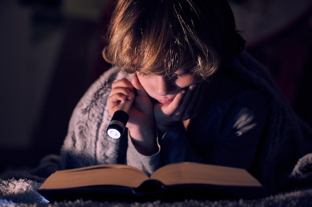Niño tranquilo leyendo un libro con una linterna encendida mientras está tendido en la cama bajo una manta en una habitación oscura por la noche en casa