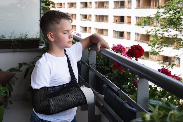 Niño triste con un brazo roto enyesado mira al patio desde el balcón