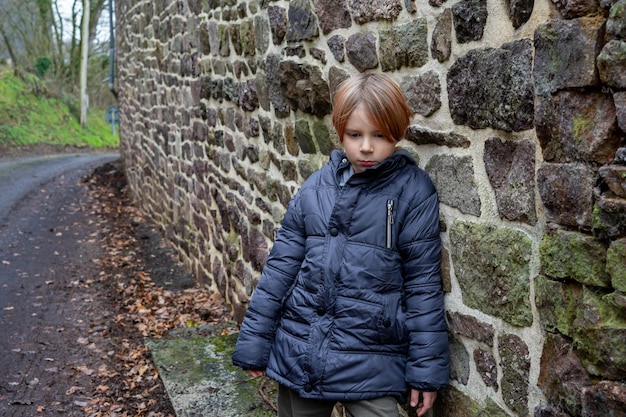 Niño triste cerca del muro de piedra Un niño de nueve años con una chaqueta abrigada se para con la cabeza inclinada Un niño que experimenta emociones negativas