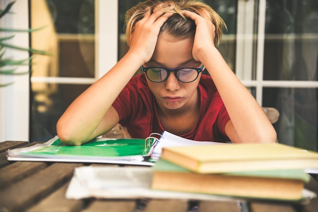 Foto niño triste con la mano en el cabello mirando libros mientras está sentado junto a la mesa