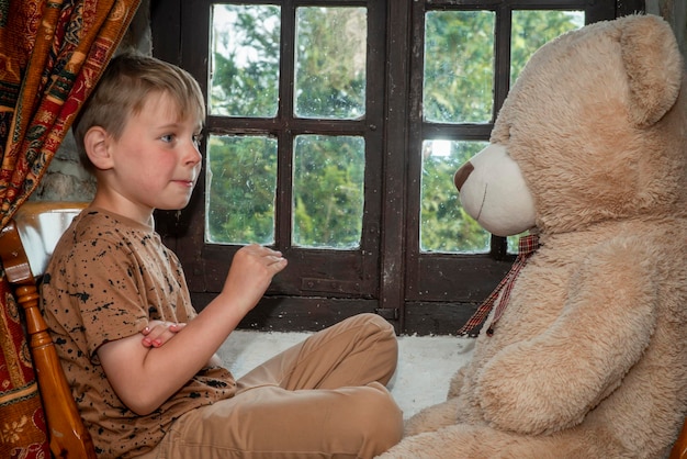 Niño triste de ocho años con un gran oso de peluche cerca de la ventana