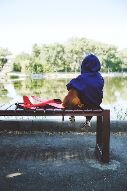 Niño triste sentado solo con un perro y un avión de juguete en un banco junto a la orilla del río diagnosticando el inicio temprano